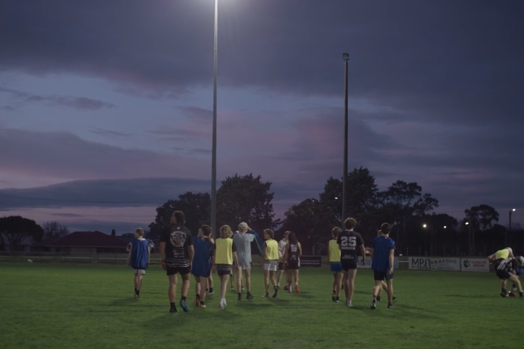 A group of around 12 young kids on a almost night setting in a rugby field playing with each other 